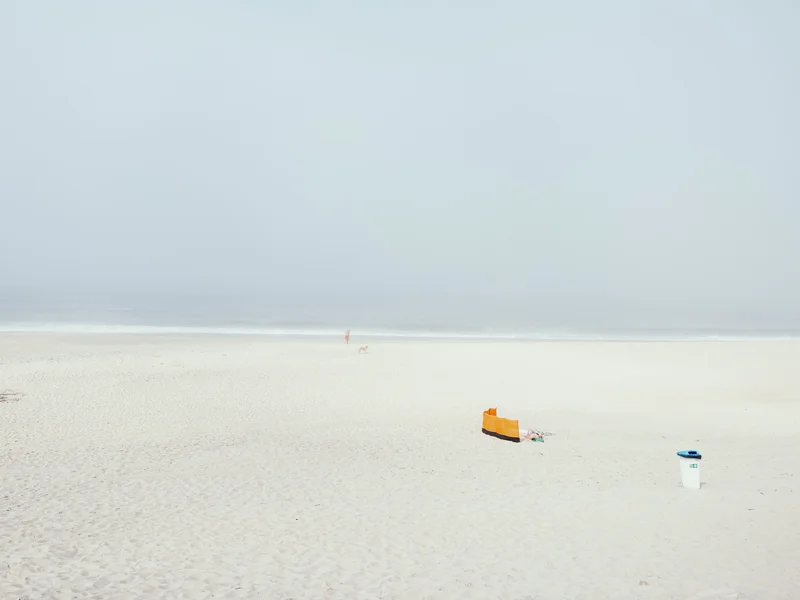 A minimalistic beach scene with a distant person and dog near the shoreline, and an orange windbreaker with a trash bin in the foreground.