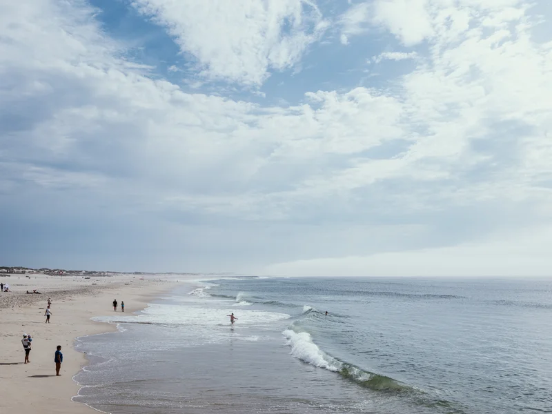 A sandy beach with people walking along the shore under a cloudy sky.