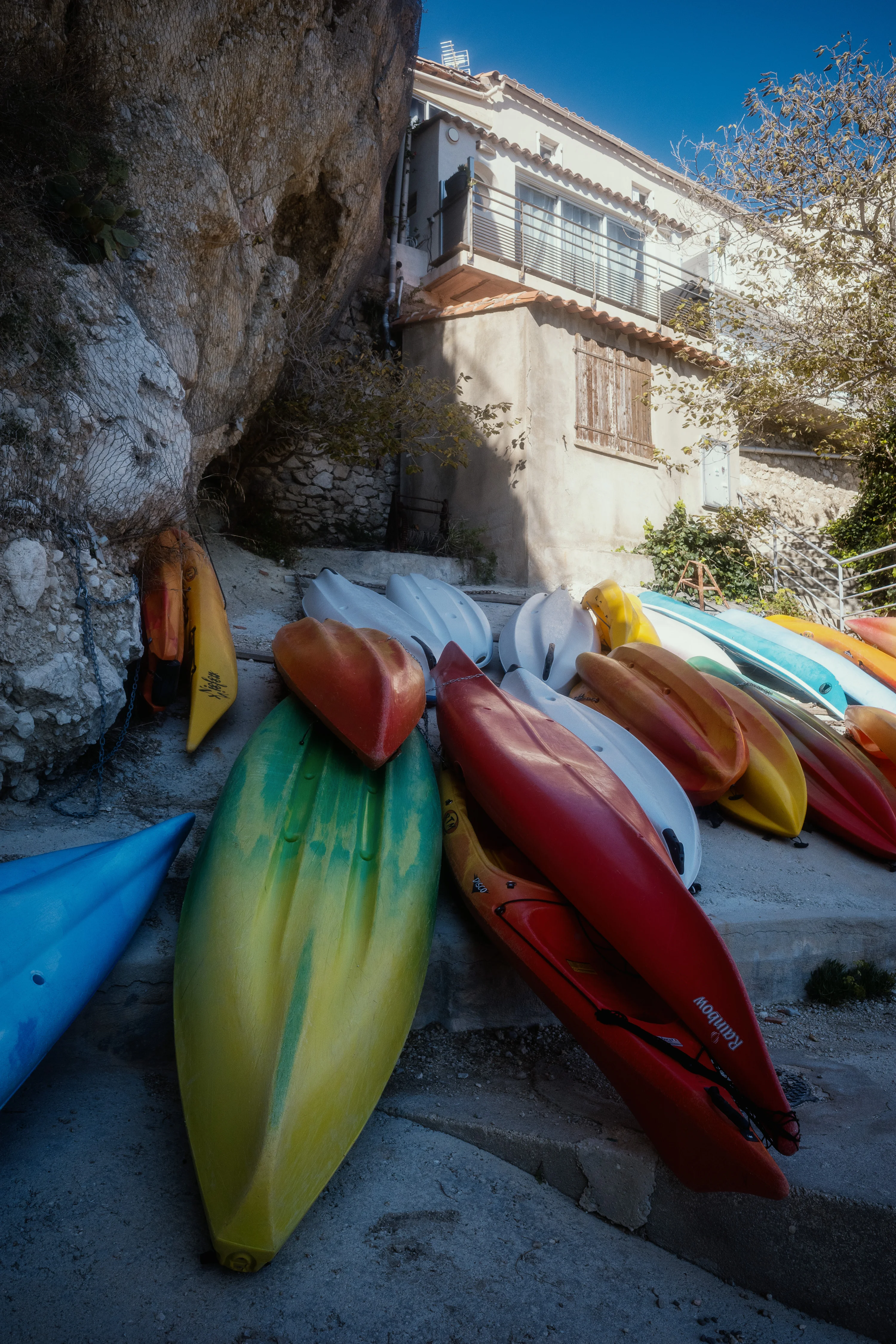 Several colorful kayaks stacked on a rocky shore beside a weathered building.