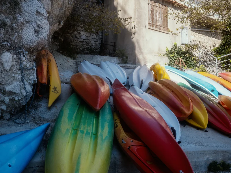 Several colorful kayaks stacked on a rocky shore beside a weathered building.