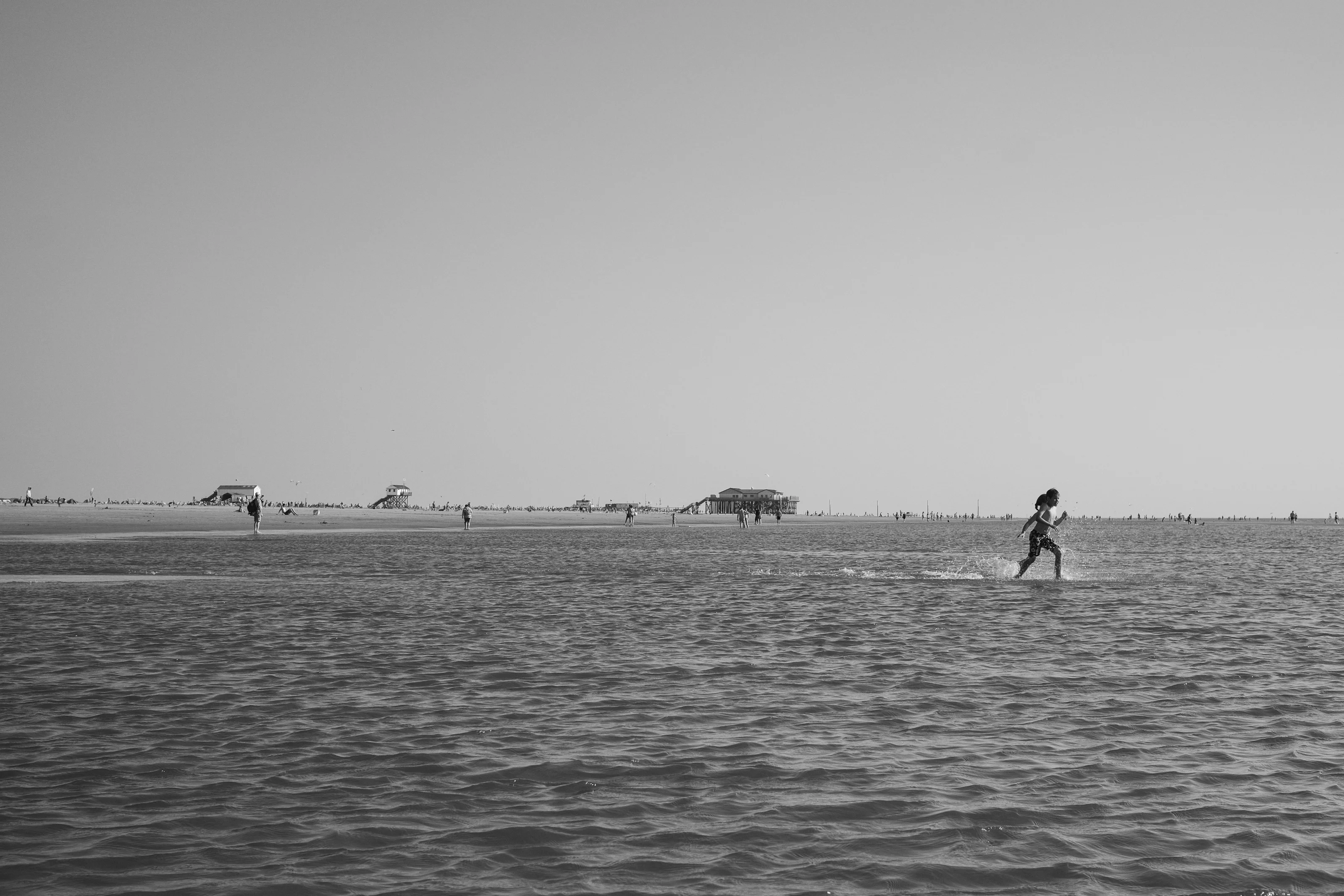 A person running through shallow water on a beach with distant structures and people in the background.