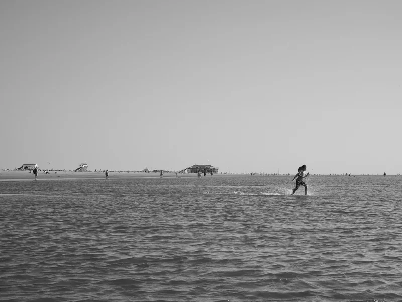 A person running through shallow water on a beach with distant structures and people in the background.