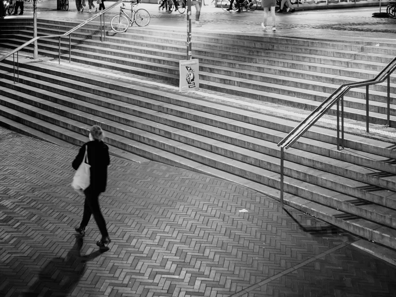 A person walks across a herringbone brick pattern with stairs and railings in the background.
