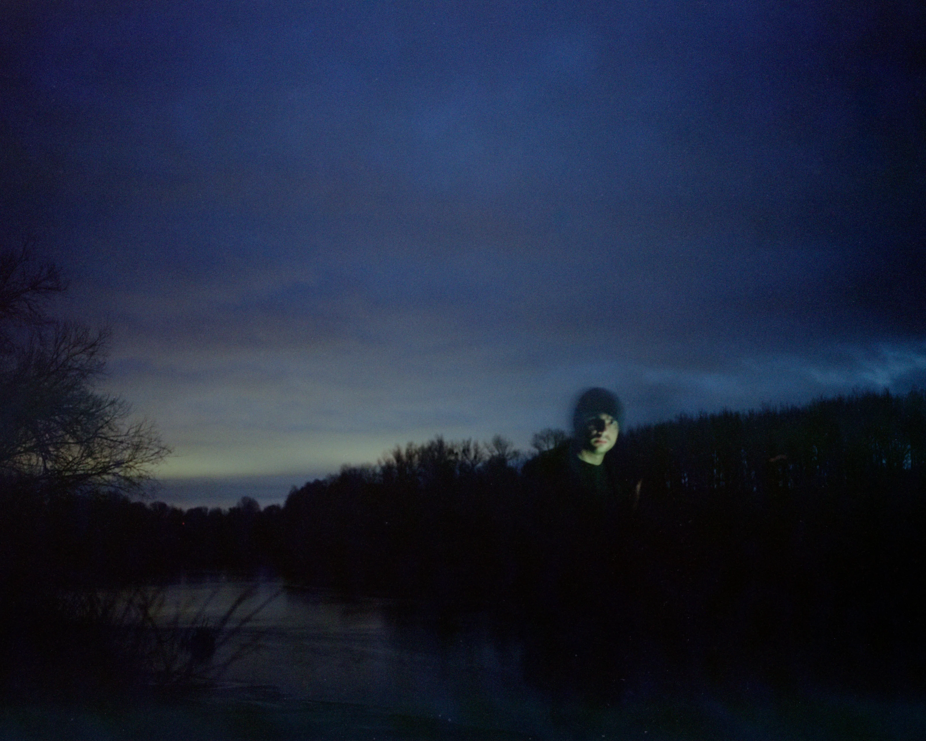 A person with a faintly illuminated face stands in front of a dark, twilight landscape with trees and water.