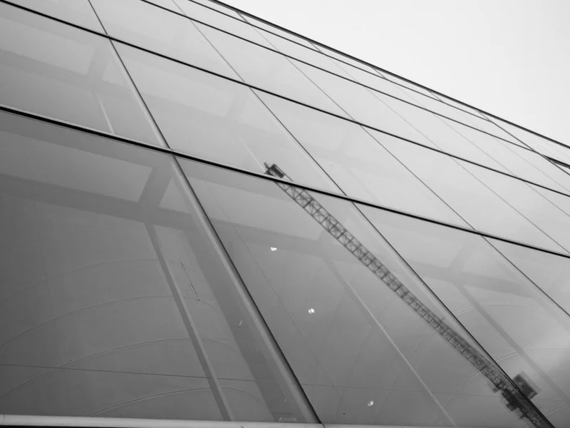 Black and white image of a modern glass building with a crane reflected on the facade.