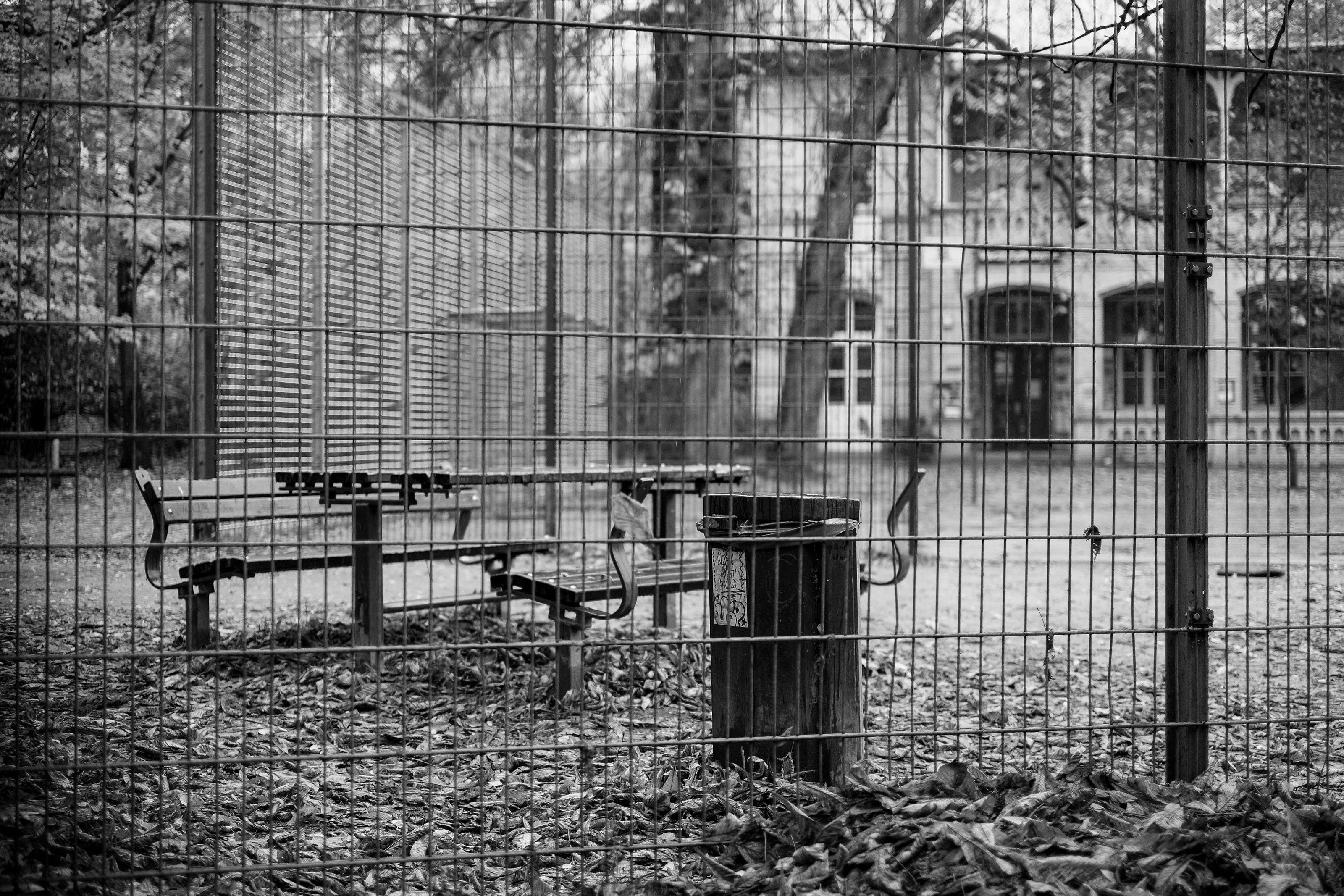 Black and white photo of an outdoor area with benches and a trash can behind a metal fence.