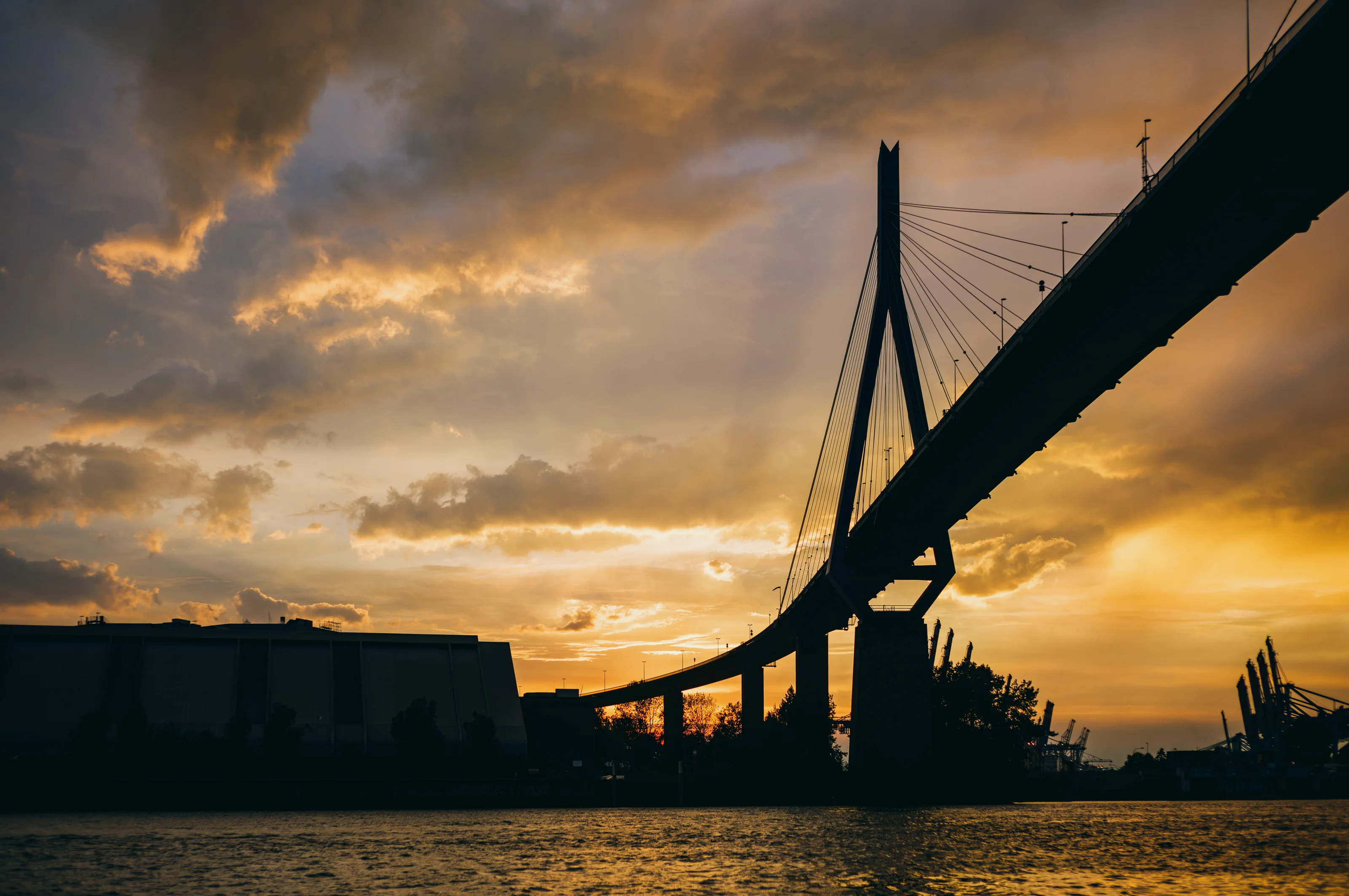 Silhouette of a suspension bridge at sunset with dramatic clouds and orange sky.
