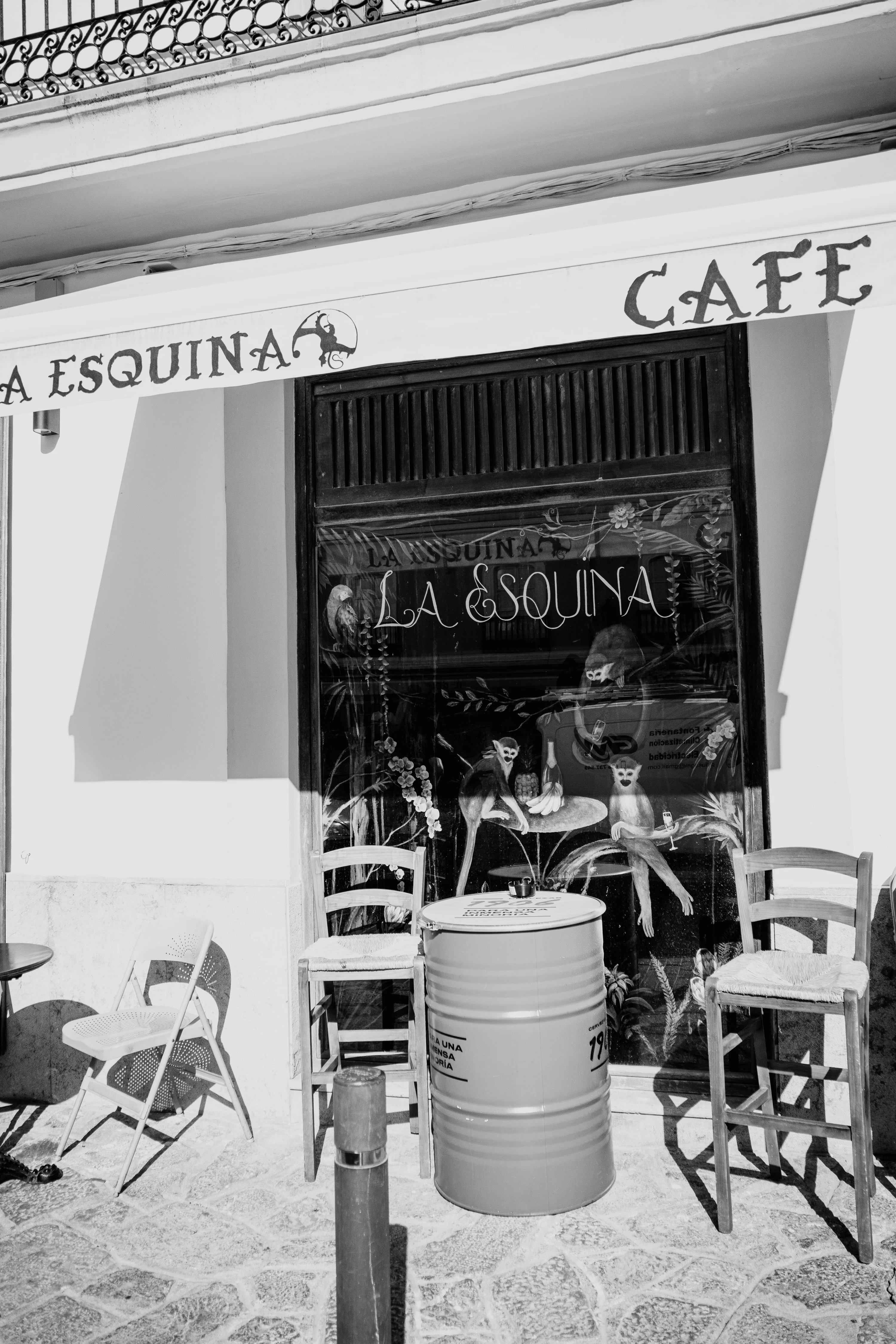 Black and white photo of a cafe entrance with chairs and a barrel outside.