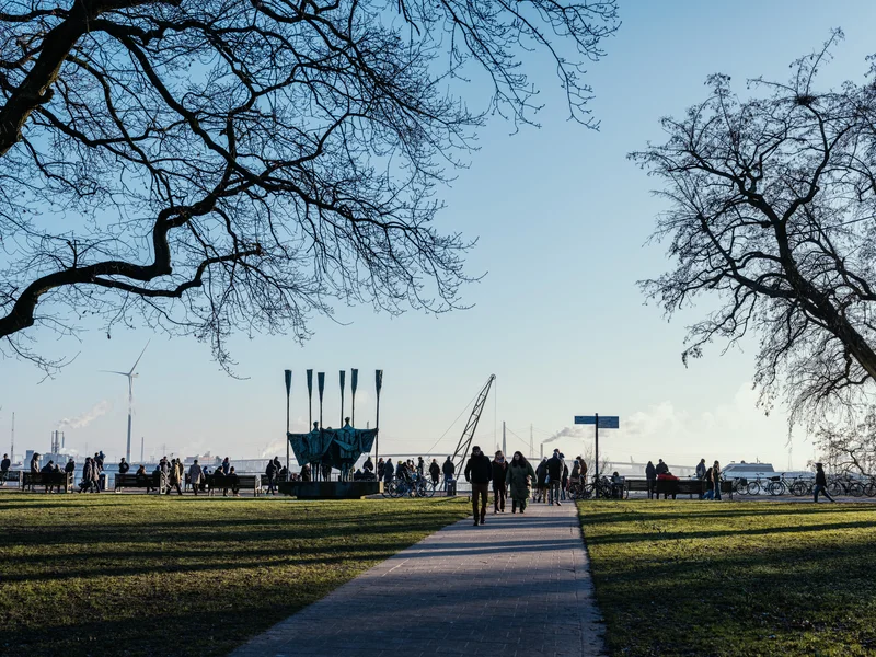 People walking in a park with leafless trees and an industrial backdrop.