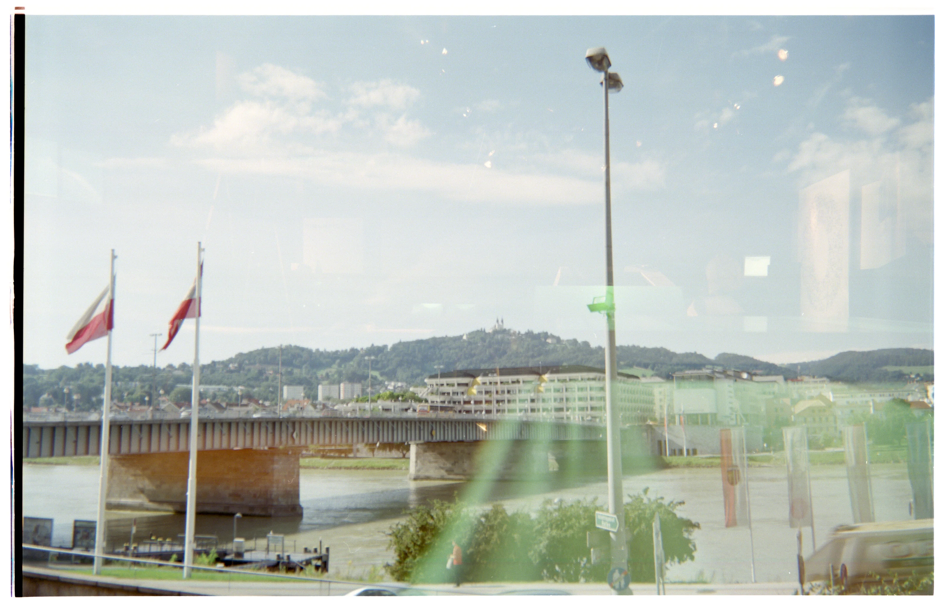 Cityscape with a bridge over a river, flags, and a hillside in the background.