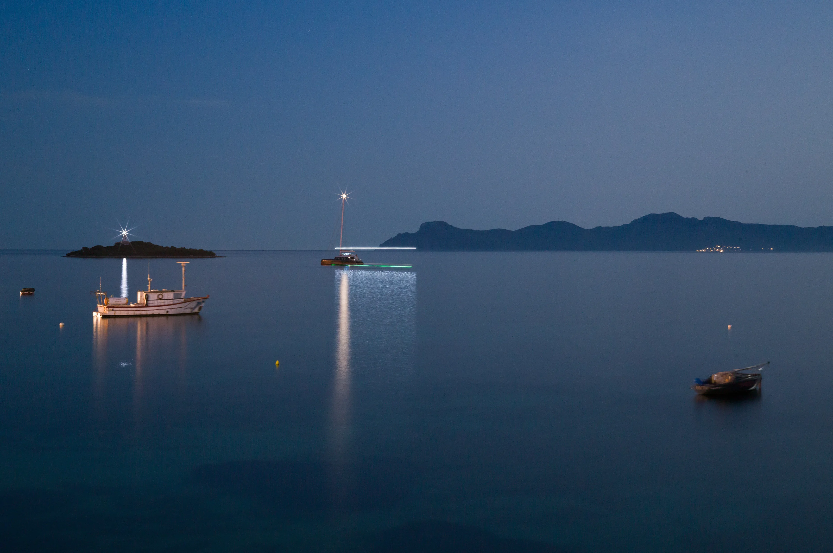 Boats are illuminated on calm waters at dusk with a distant island in the background.