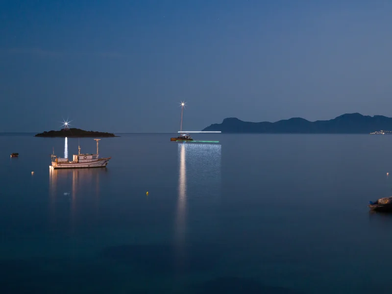 Boats are illuminated on calm waters at dusk with a distant island in the background.