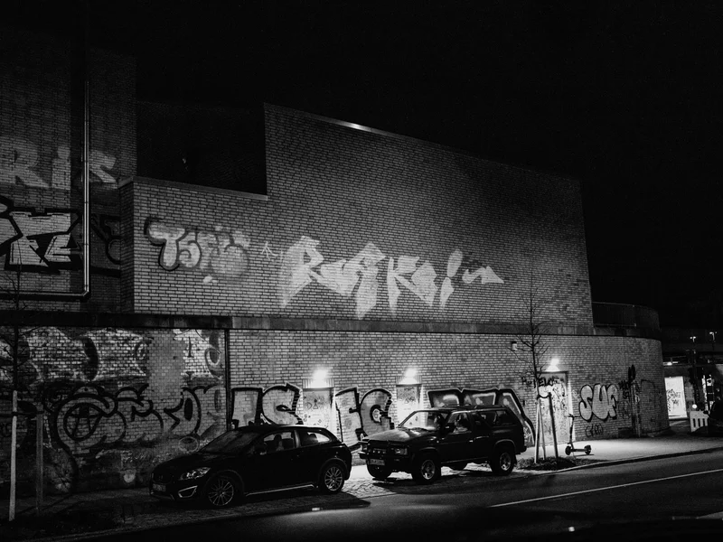 Black and white photo of a brick wall with graffiti and parked cars on a dimly lit street at night.