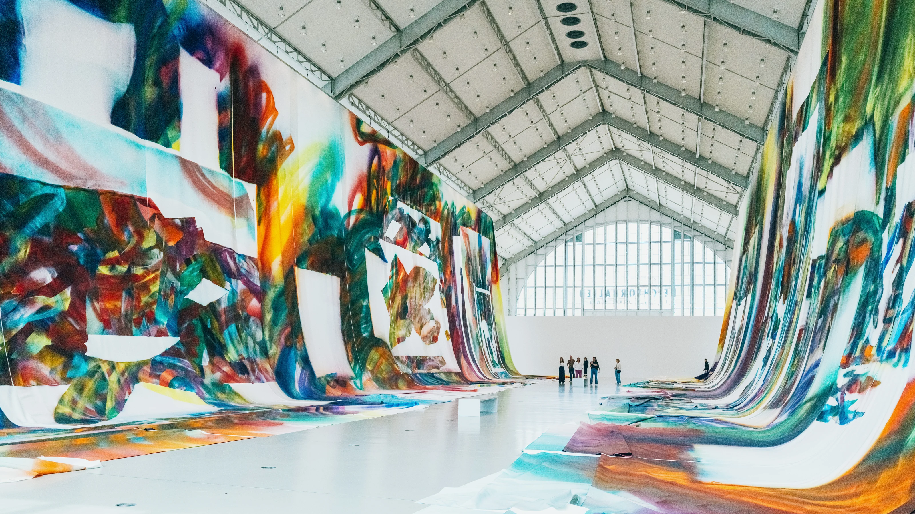 Colorful abstract murals hang on both sides of a spacious gallery with people observing from a distance.