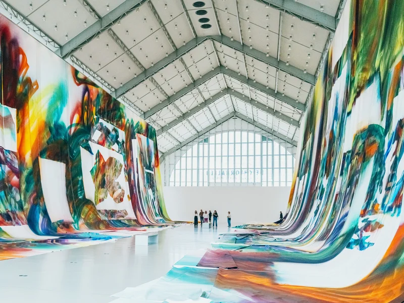 Colorful abstract murals hang on both sides of a spacious gallery with people observing from a distance.