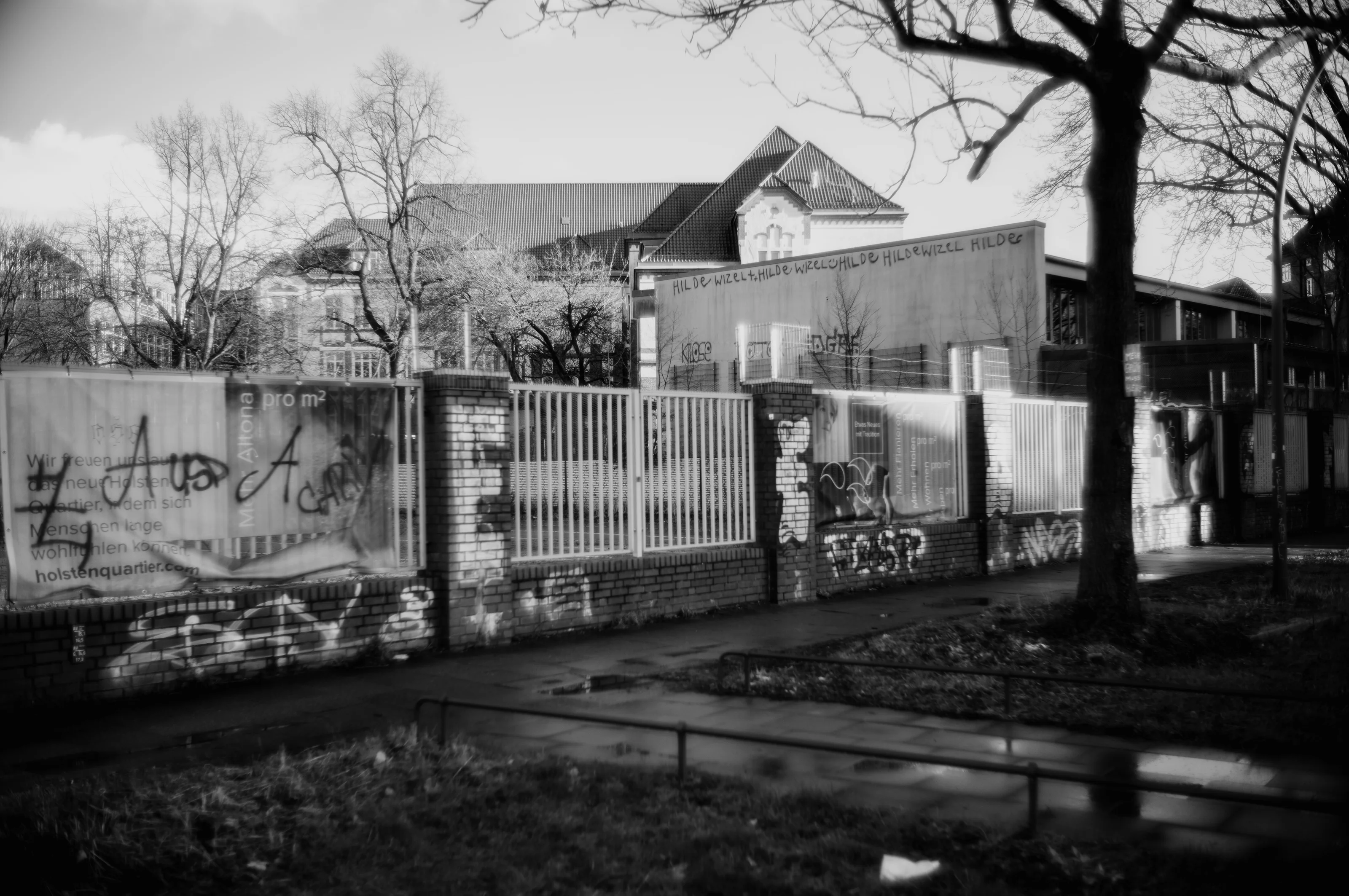 A graffiti-covered brick wall with metal fencing and a building in the background on a cloudy day.