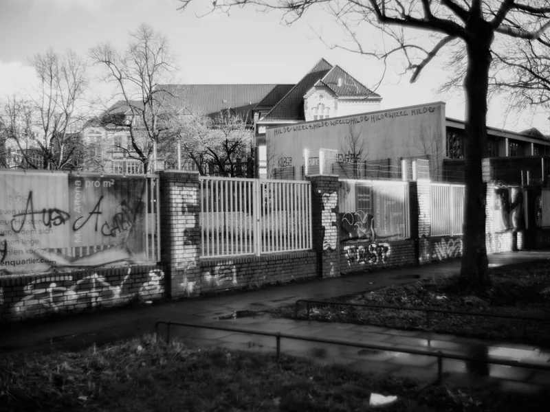 A graffiti-covered brick wall with metal fencing and a building in the background on a cloudy day.