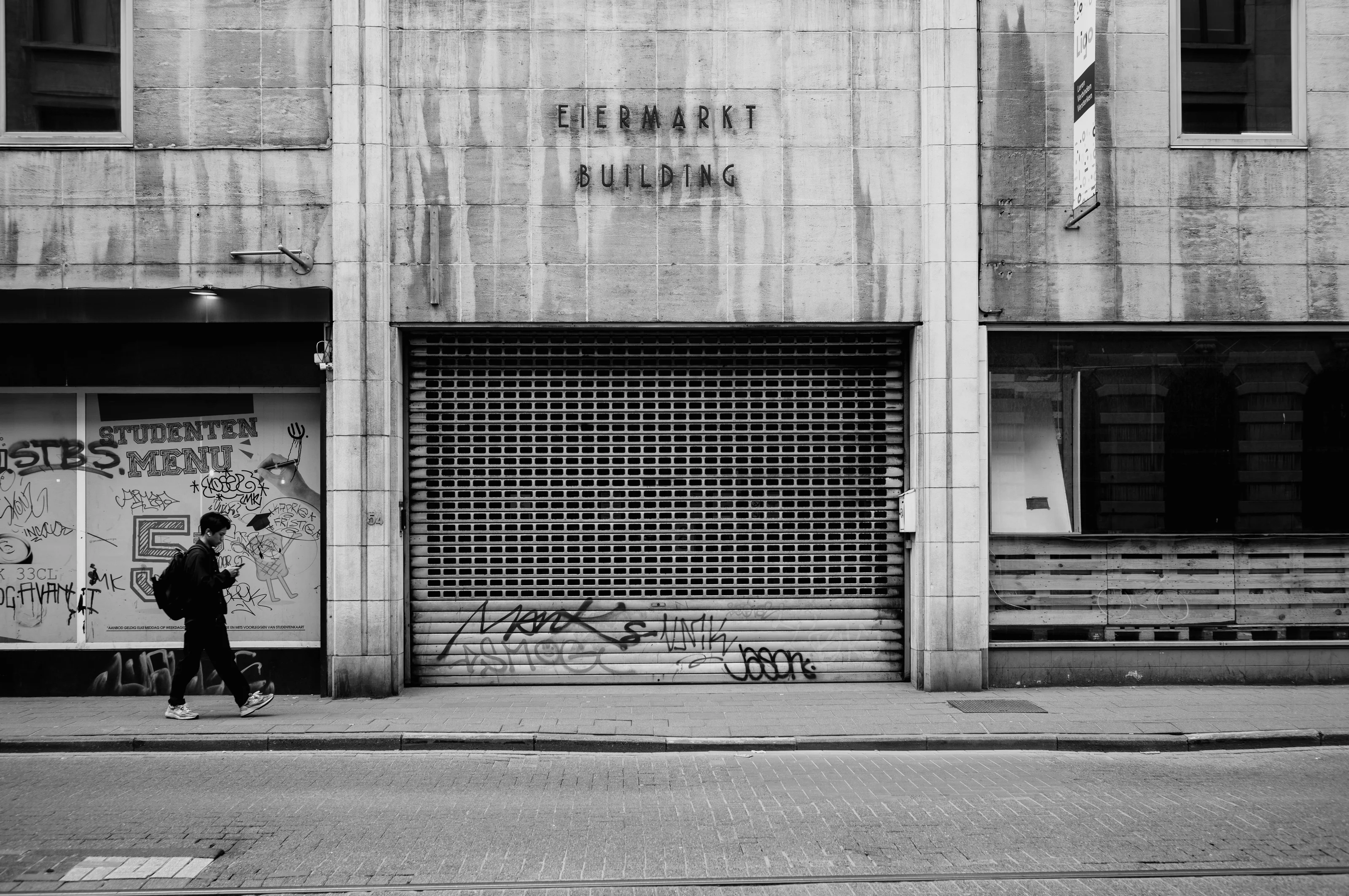 A person walks past a building with a closed shutter and graffiti.