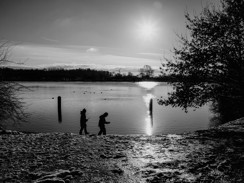 Two people walking near a lake with a bright sun reflecting on the water.