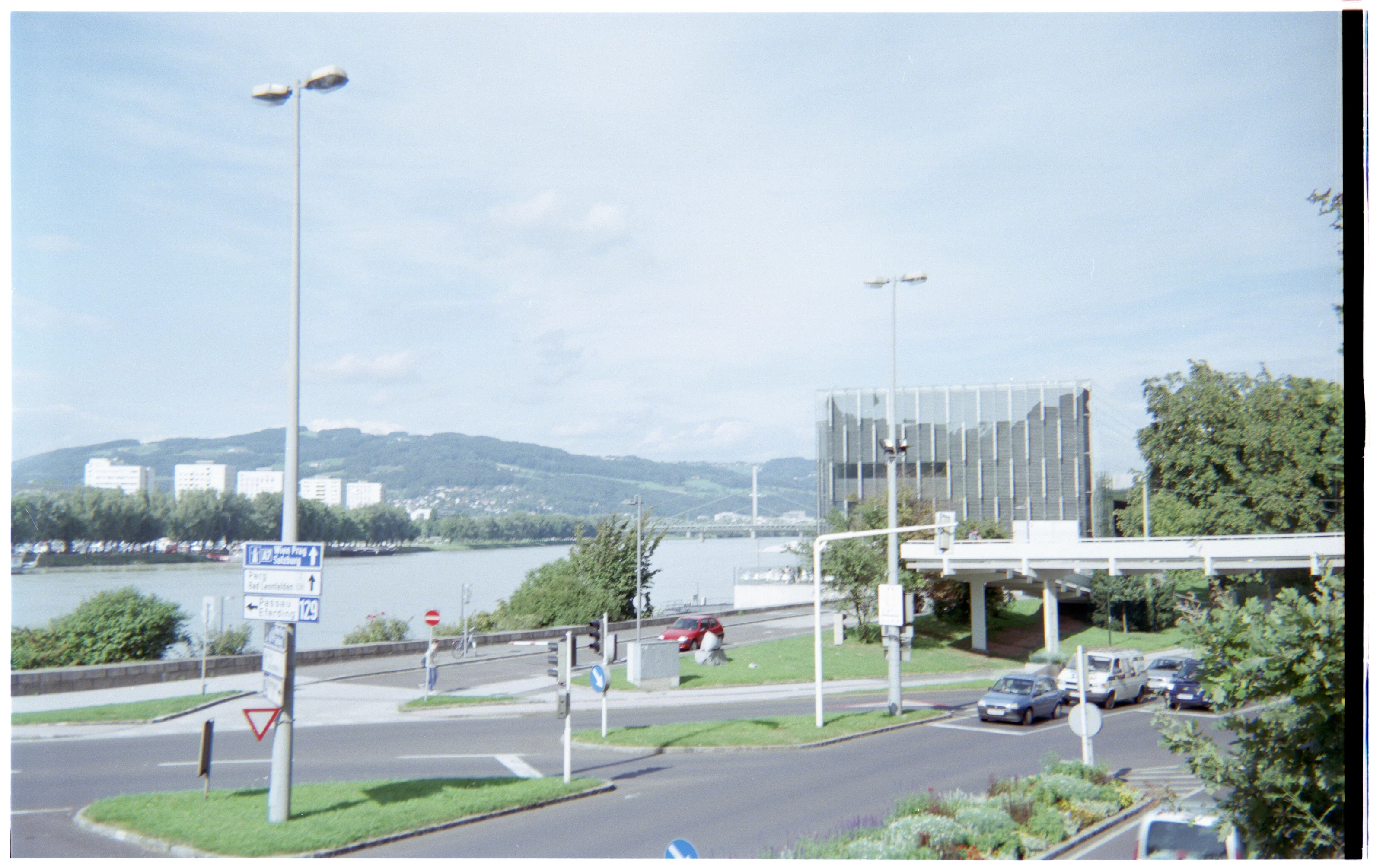 A riverside urban scene with buildings, a bridge, and street signs under a blue sky.