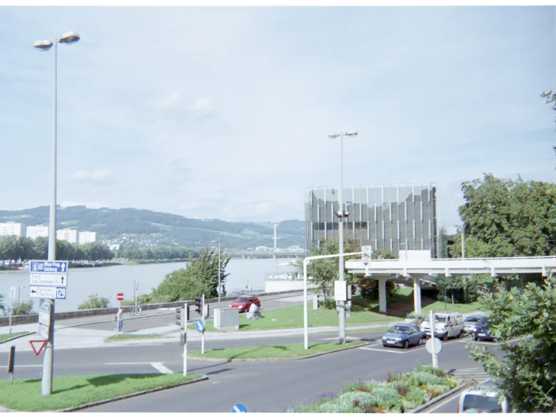 A riverside urban scene with buildings, a bridge, and street signs under a blue sky.