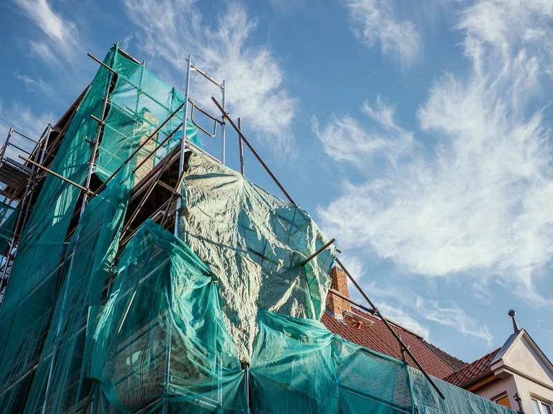 Building under construction covered with green netting against a blue sky with wispy clouds.