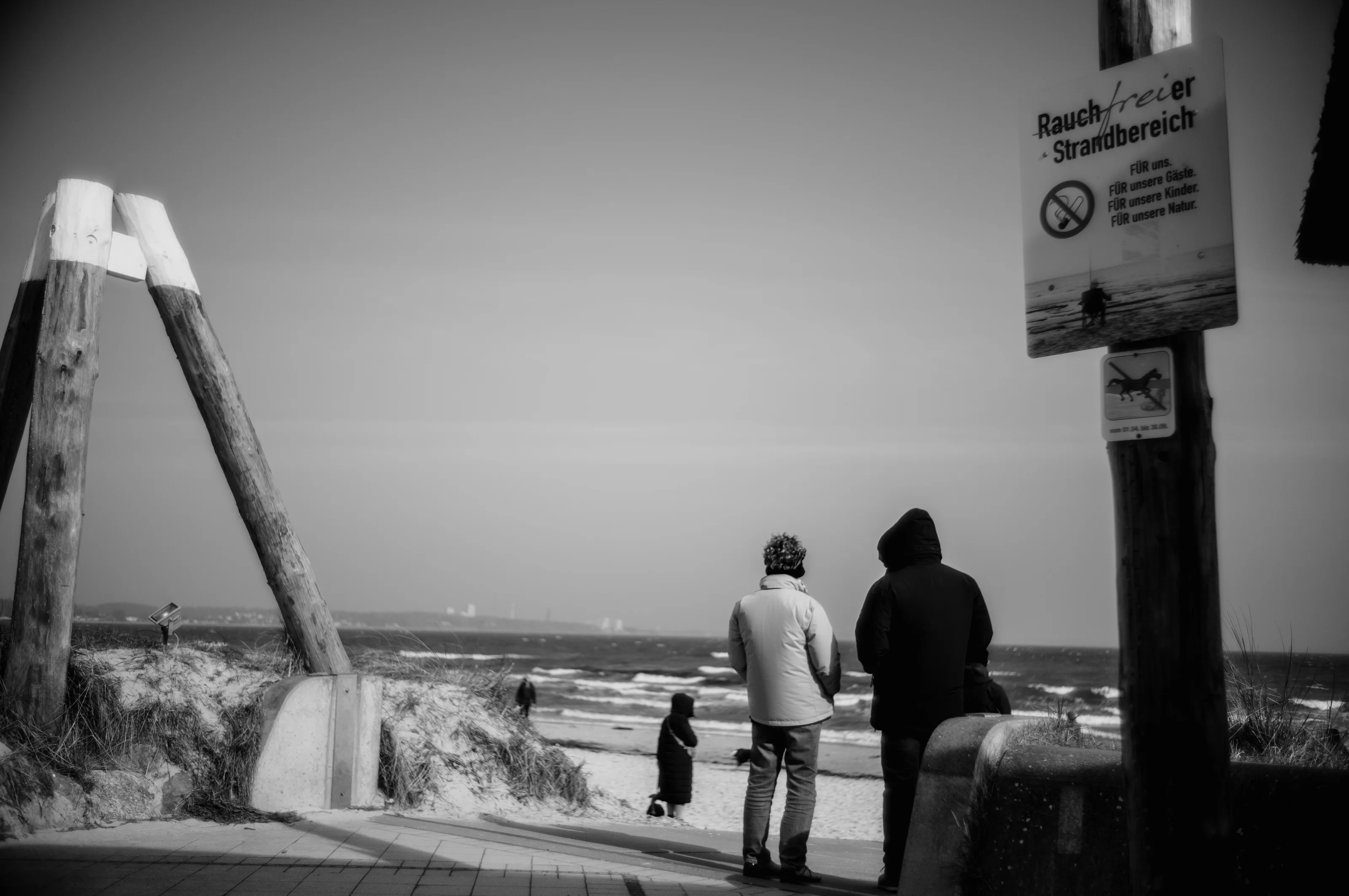 Two people in jackets stand on a beach walkway under a no smoking sign with the ocean in the background.