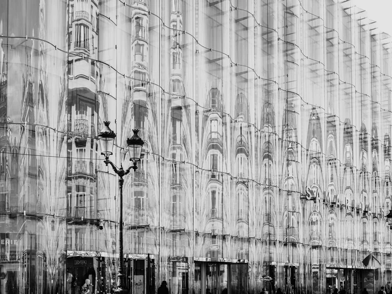 Black and white photo of a street scene with a modern glass building reflecting older architecture.