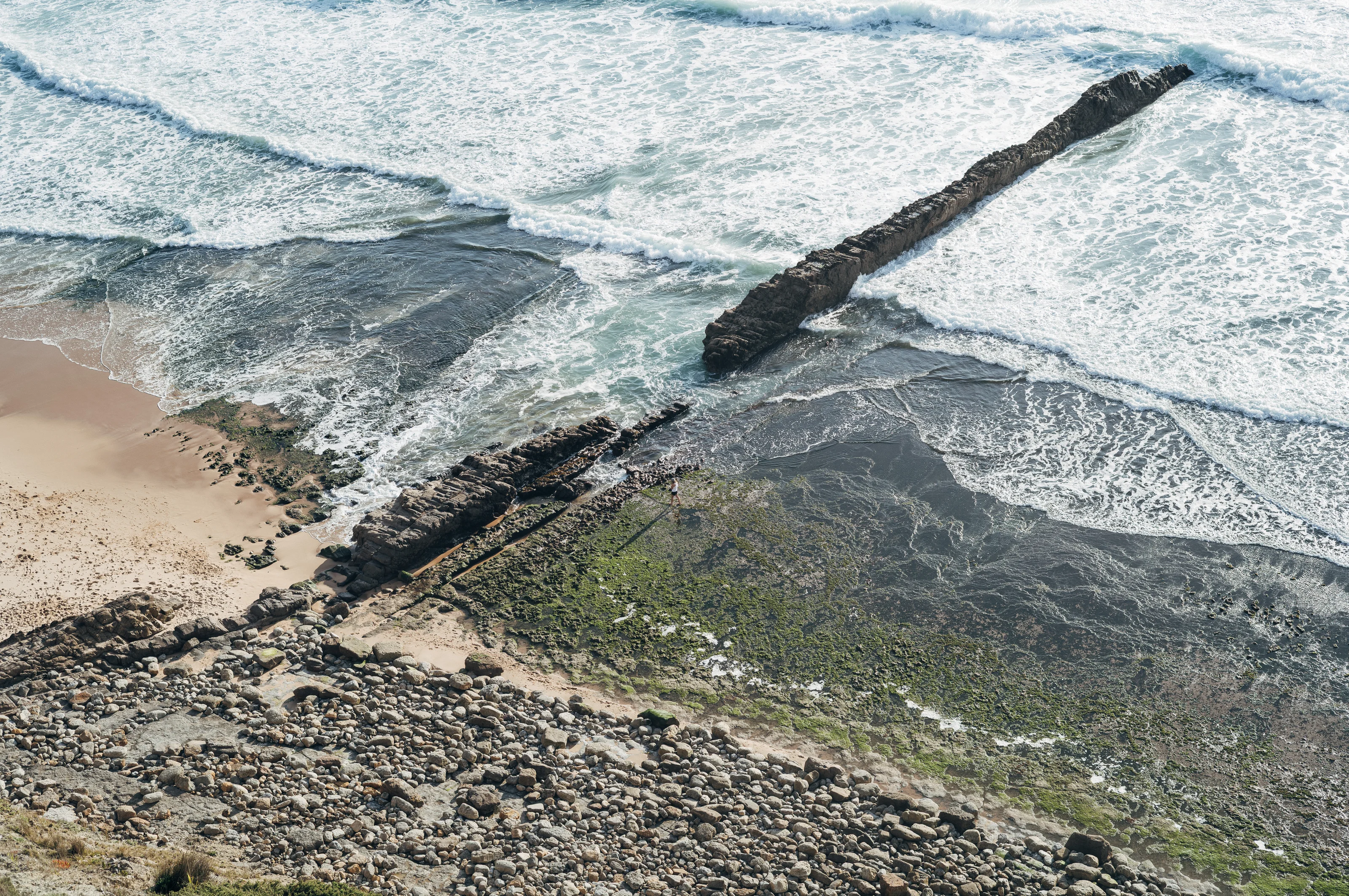 Coastal scene with waves crashing against a rocky shore and a long rock formation extending into the ocean.