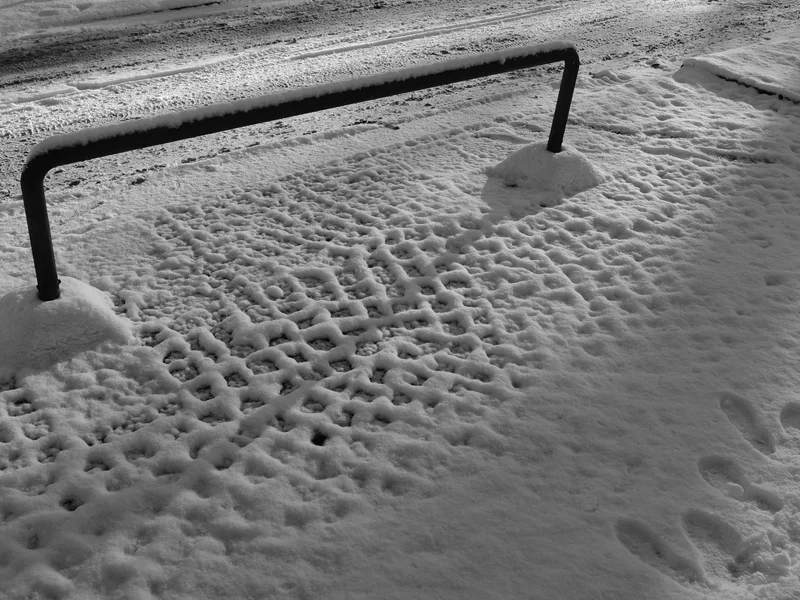 Snow-covered ground with tire and footprint impressions alongside a metal barrier.