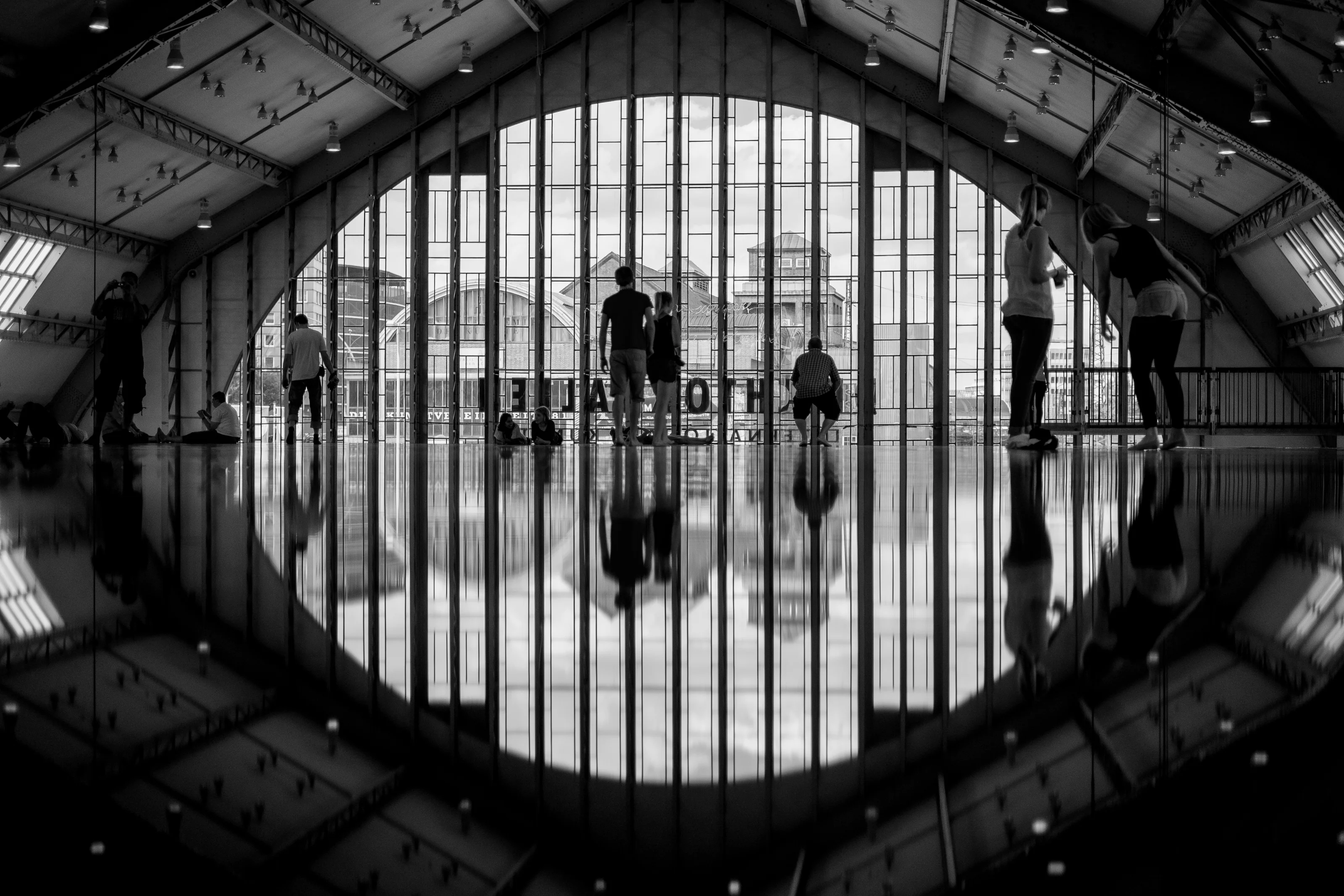 Silhouetted people in an indoor space with floor reflections and large windows showing a cityscape.