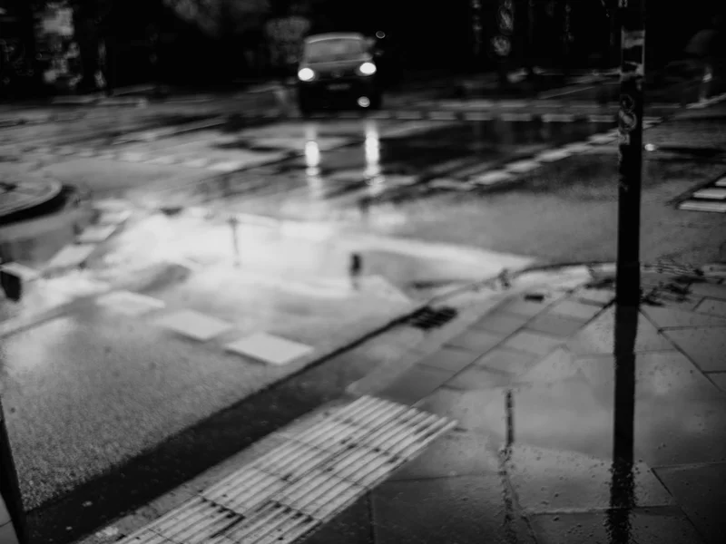 A blurry black and white street scene with a car approaching and reflections on wet pavement.