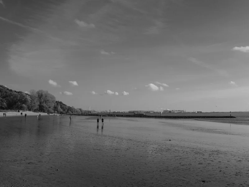 Black and white photo of a beach with people walking along the shore and a distant industrial skyline.