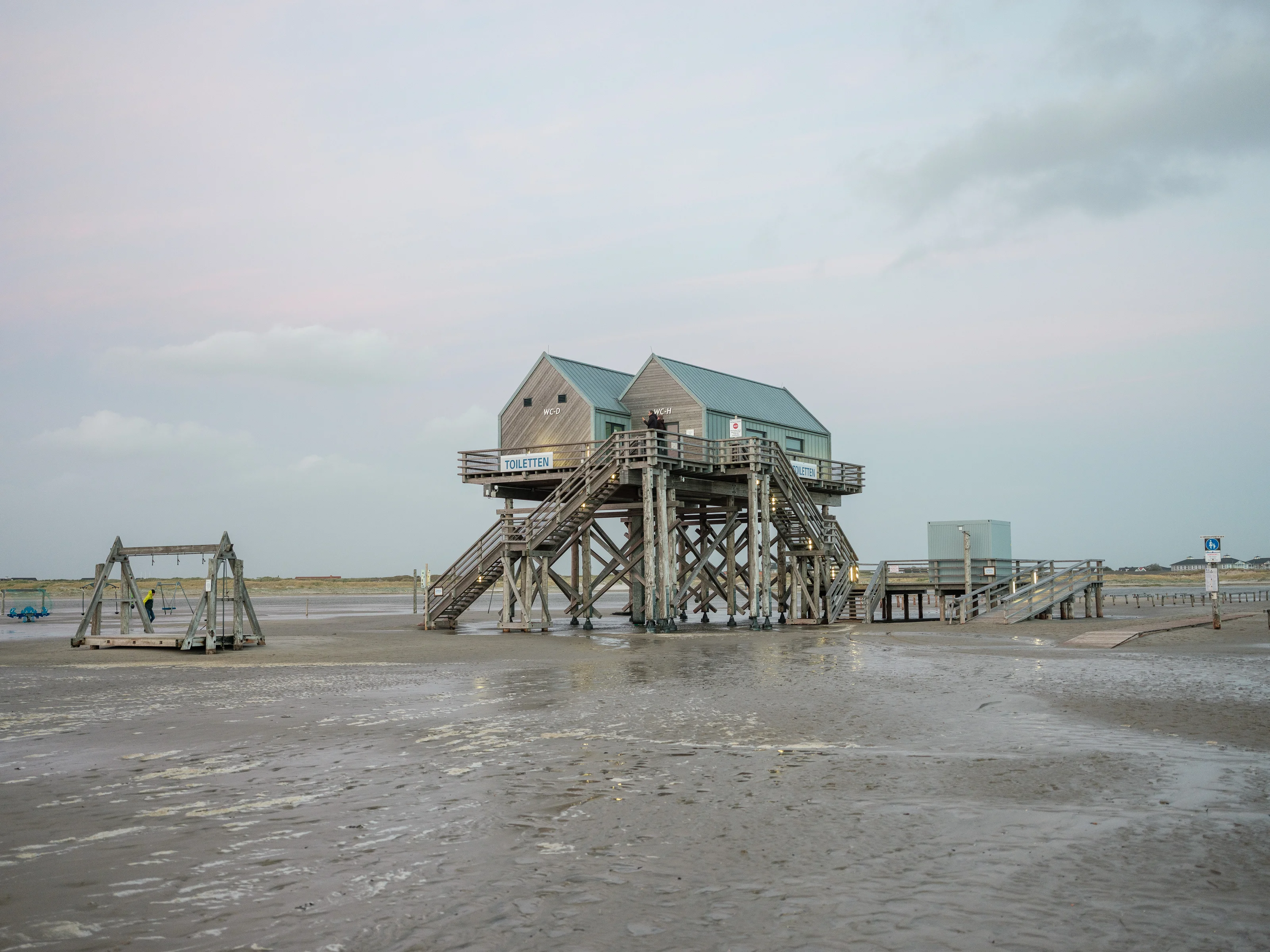 Elevated wooden building on stilts at a sandy beach with a swing structure nearby.