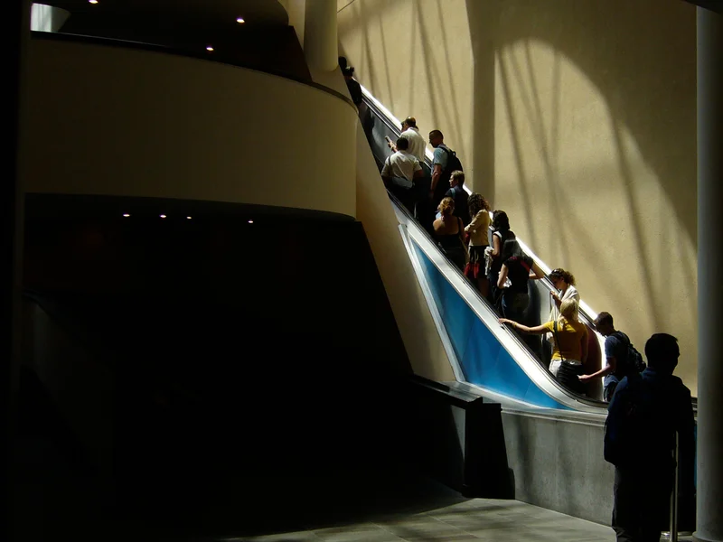 People riding an upward escalator in a building with sunlight casting shadows.