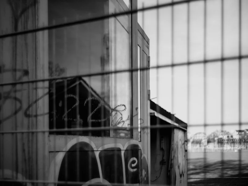 A monochrome photograph of a building with graffiti, viewed through a wire fence.