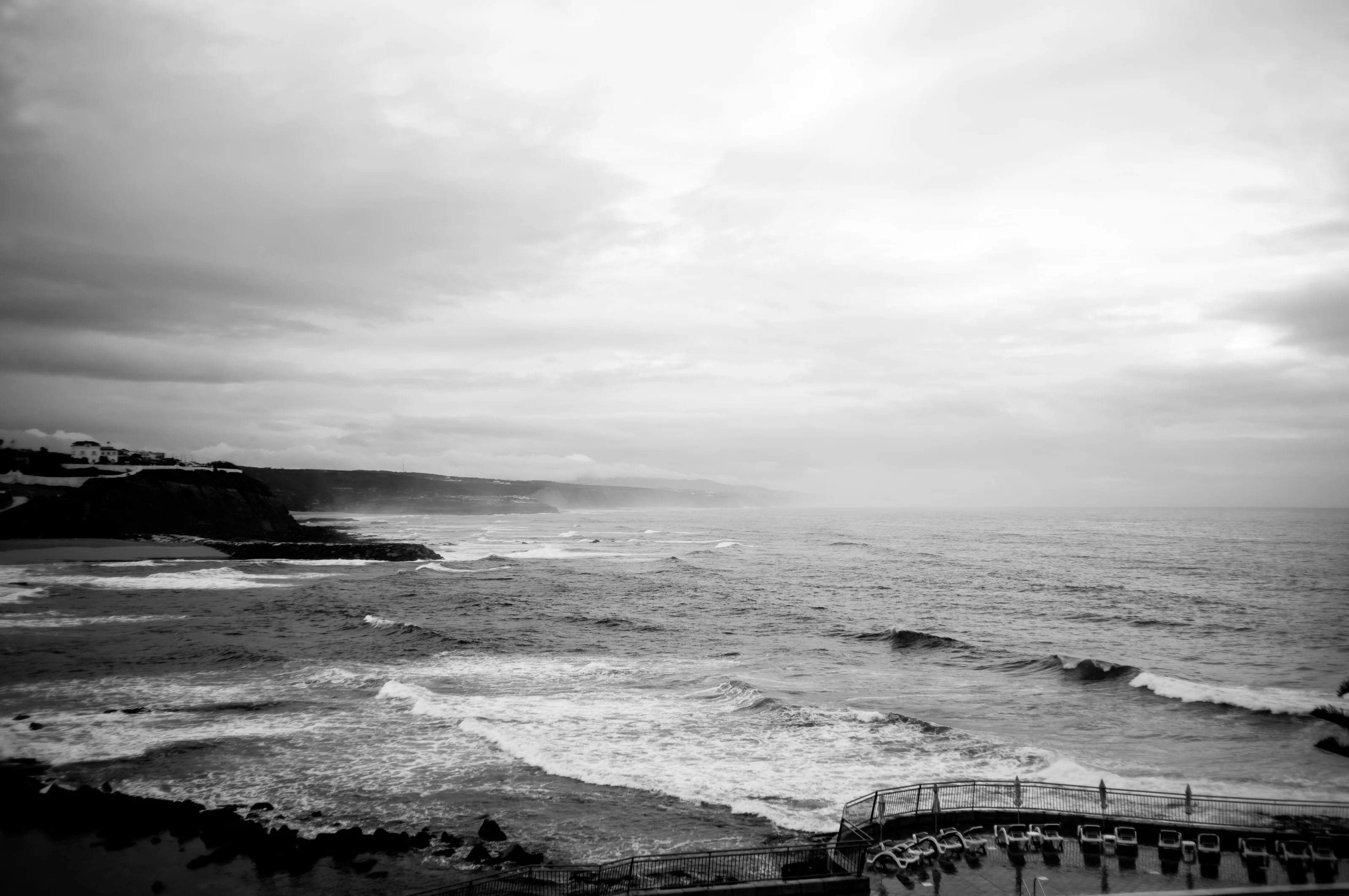 Black and white seascape with crashing waves, rocky shoreline, and a fenced lookout.