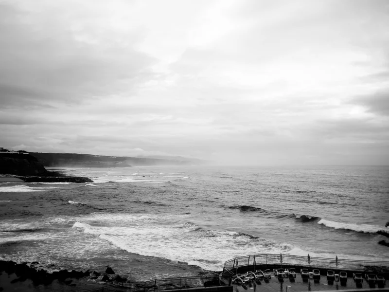 Black and white seascape with crashing waves, rocky shoreline, and a fenced lookout.