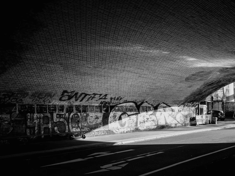 Black and white photo of a graffitied brick underpass with sunlight streaming in.