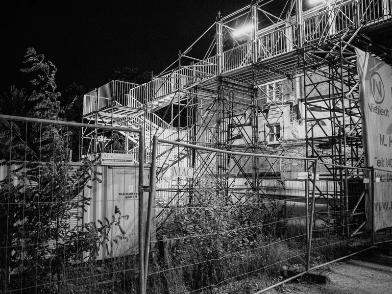 A black and white photograph of a construction site with scaffolding and lighting.