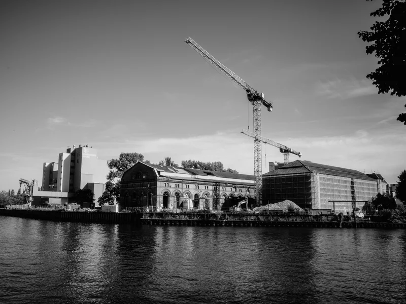 A black and white photograph of a riverside industrial site with cranes and large buildings.